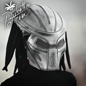K&ouml;nnte beinhalten: Eine silberne Predator-Maske aus Leder mit schwarzen Dreadlocks. Die Maske hat ein detailliertes Design mit gen&auml;hten Linien und einem Netz-Augenabdeckung.
