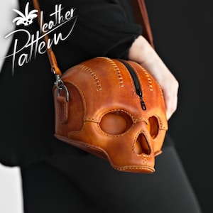 K&ouml;nnte beinhalten: Eine beigefarbene Lederschultertasche in Form eines menschlichen Sch&auml;dels mit sichtbaren N&auml;hten entlang der Kanten und einem schwarzen Rei&szlig;verschluss an der Oberseite. Die Tasche hat Aussparungen f&uuml;r Augen und Nase sowie einen d&uuml;nnen Lederriemen.