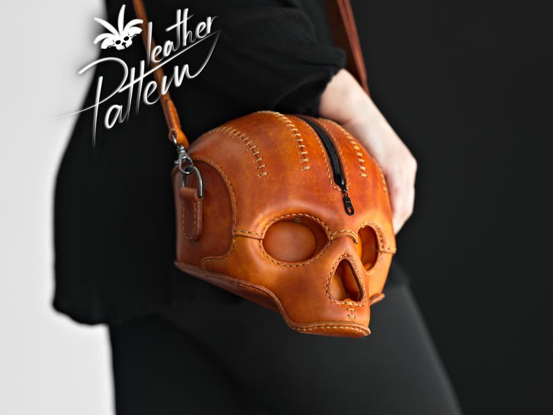 K&ouml;nnte beinhalten: Eine beigefarbene Lederschultertasche in Form eines menschlichen Sch&auml;dels mit sichtbaren N&auml;hten entlang der Kanten und einem schwarzen Rei&szlig;verschluss an der Oberseite. Die Tasche hat Aussparungen f&uuml;r Augen und Nase sowie einen d&uuml;nnen Lederriemen.