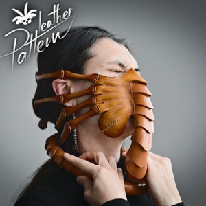 K&ouml;nnte beinhalten: Eine braune Ledermaske mit einem Facehugger-Design. Die Maske hat einen langen, spitzen R&uuml;ssel und mehrere Arme, die sich an den Seiten erstrecken. Die Maske wird von einer Person mit geschlossenen Augen getragen.