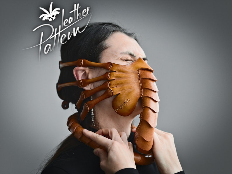K&ouml;nnte beinhalten: Eine braune Ledermaske mit einem Facehugger-Design. Die Maske hat einen langen, spitzen R&uuml;ssel und mehrere Arme, die sich an den Seiten erstrecken. Die Maske wird von einer Person mit geschlossenen Augen getragen.