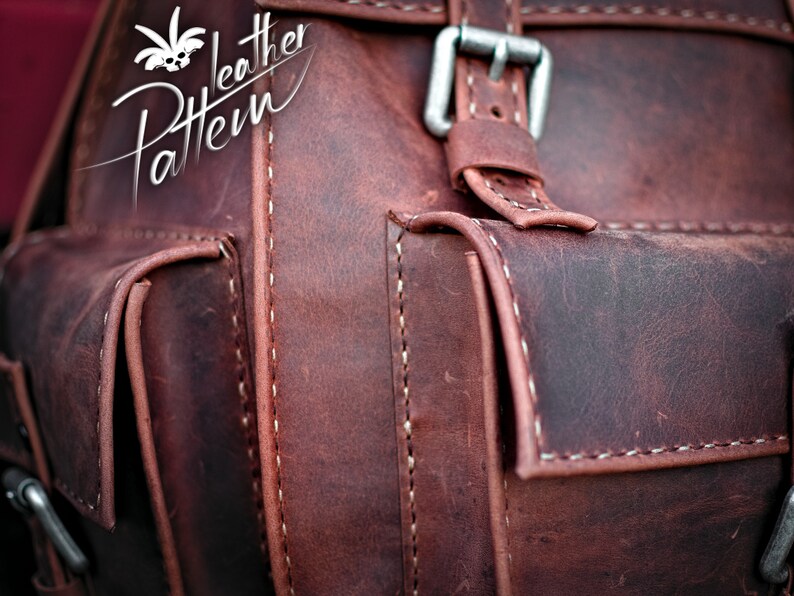 May include: Close-up of a brown leather backpack with a silver buckle and white stitching. The backpack has a flap pocket with a smaller pocket underneath.