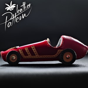 K&ouml;nnte beinhalten: Ein handgefertigtes rotes Leder-Spielzeug-Rennauto mit braunen Akzenten und schwarzen R&auml;dern. Das Auto hat dekorative N&auml;hte und den Text "Leather Pattern" in wei&szlig;er Schrift. Das Spielzeugauto ist ein Design im Vintage-Stil.