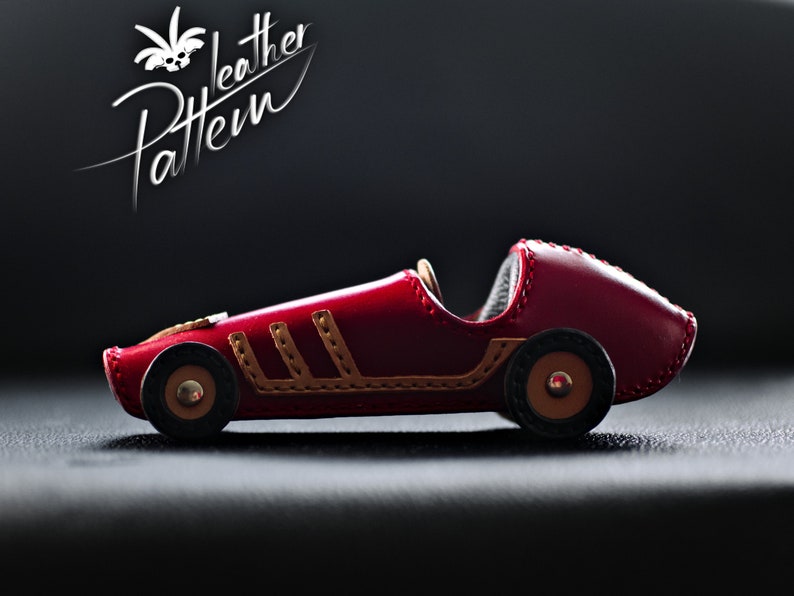 K&ouml;nnte beinhalten: Ein handgefertigtes rotes Leder-Spielzeug-Rennauto mit braunen Akzenten und schwarzen R&auml;dern. Das Auto hat dekorative N&auml;hte und den Text "Leather Pattern" in wei&szlig;er Schrift. Das Spielzeugauto ist ein Design im Vintage-Stil.