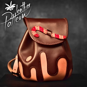 May include: A brown leather backpack with a candy cane design. The backpack has a flap with a buckle closure and a small pocket on the front. The candy cane is made of red and white leather and is sewn onto the flap.