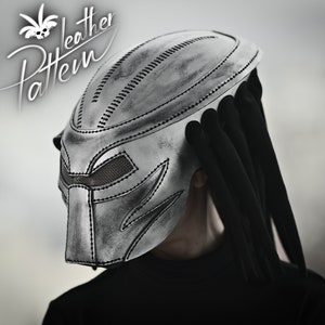 K&ouml;nnte beinhalten: Ein silberner Predator-Helm mit einer schwarzen Stoffverl&auml;ngerung, die an Dreadlocks erinnert. Der Helm hat ein detailliertes Design mit gen&auml;hten Linien und einem Mesh-Visier.
