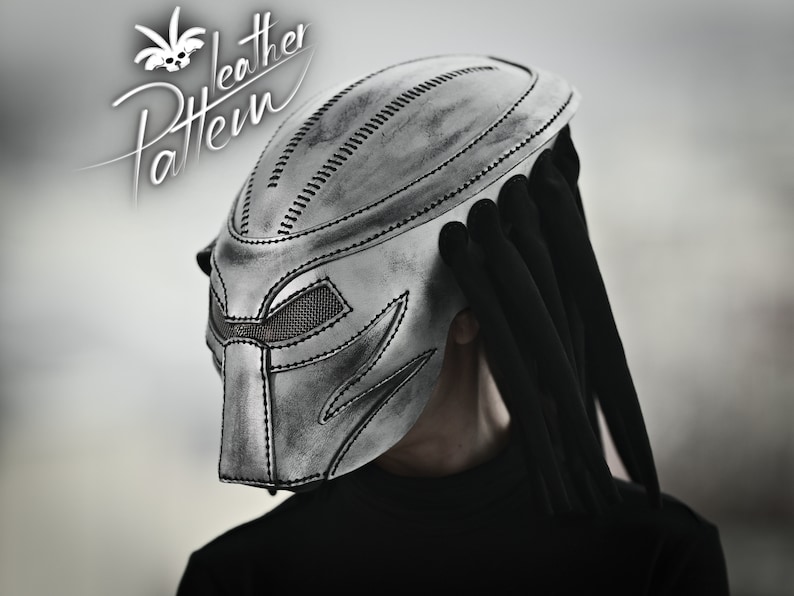 K&ouml;nnte beinhalten: Ein silberner Predator-Helm mit einer schwarzen Stoffverl&auml;ngerung, die an Dreadlocks erinnert. Der Helm hat ein detailliertes Design mit gen&auml;hten Linien und einem Mesh-Visier.