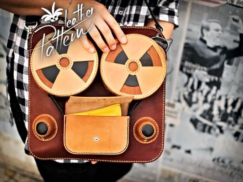K&ouml;nnte beinhalten: Eine braune Ledertasche mit zwei runden Taschen, die mit einem schwarzen und braunen Design verziert sind. Die Tasche hat eine Klappe mit einer kleinen Tasche und einem gelben Stoff im Inneren. Die Tasche hat einen Riemen und der Text "Leather Pattern" ist auf der Vorderseite aufgedruckt.