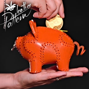 May include: A bright orange leather piggy bank with a stitched design. A hand is placing a gold coin into the slot.