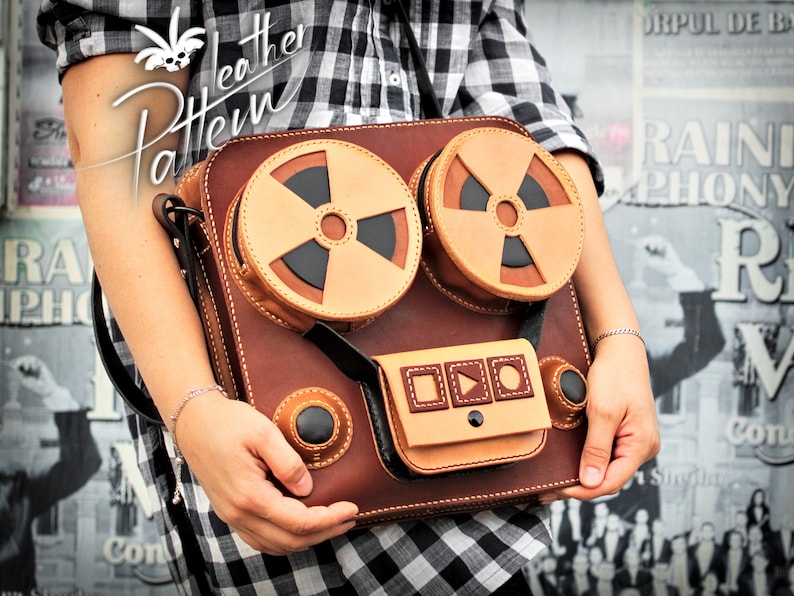 K&ouml;nnte beinhalten: Eine braune Ledertasche in Form eines Vintage-Tonbandger&auml;ts. Die Tasche hat zwei gro&szlig;e Spulen, ein Bedienfeld mit Kn&ouml;pfen und einen Riemen.