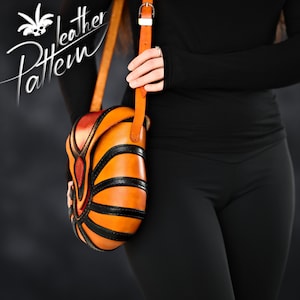 Könnte beinhalten: Eine braune Leder-Umhängetasche mit schwarzen und orangefarbenen Akzenten. Die Tasche hat ein einzigartiges geometrisches Design und einen langen Riemen.