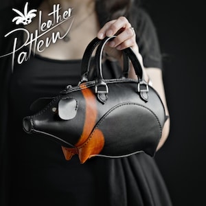 Puede incluir: Bolso de mano de cuero negro con forma de cerdo, con detalles en naranja y marrón. El bolso tiene un asa superior y un cuerpo pequeño y redondo. La imagen incluye el texto "Leather Pattern".