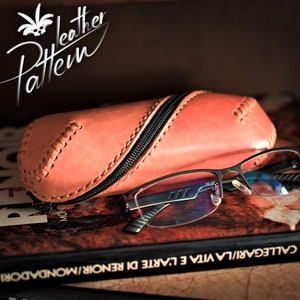 Puede incluir: Un estuche para gafas de cuero rosa con cierre de cremallera y costuras decorativas. El estuche está apoyado en un libro con el texto "PIERA CALLEGARI/LA VITA E L'ARTE DI RENOIR/MONDADORI" visible.