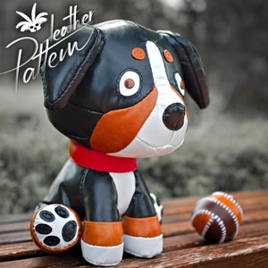 Puede incluir: Un juguete de peluche de cuero negro, marrón y blanco que representa a un perro de montaña bernés sentado en una superficie de madera con una pelota de cuero marrón a su lado. El juguete tiene un collar rojo y ojos negros.