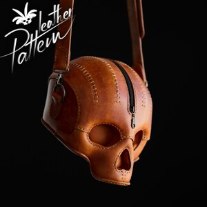 K&ouml;nnte beinhalten: Eine lohfarbene Ledertasche in Form eines menschlichen Sch&auml;dels mit sichtbaren N&auml;hten entlang der Kanten und einem schwarzen Rei&szlig;verschluss auf der Oberseite. Die Tasche hat einen langen verstellbaren Schulterriemen und den Text Pattern Leather.