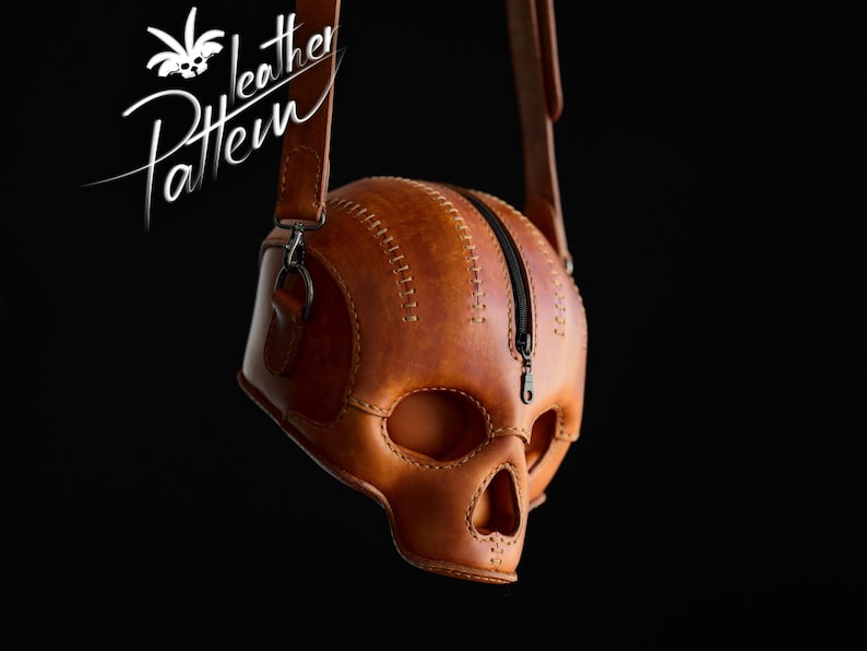 K&ouml;nnte beinhalten: Eine lohfarbene Ledertasche in Form eines menschlichen Sch&auml;dels mit sichtbaren N&auml;hten entlang der Kanten und einem schwarzen Rei&szlig;verschluss auf der Oberseite. Die Tasche hat einen langen verstellbaren Schulterriemen und den Text Pattern Leather.