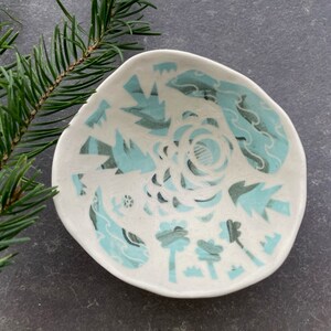May include: A small, white ceramic bowl with a teal blue and white geometric design. The bowl is slightly irregular in shape and has a rounded bottom.