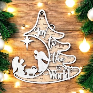 May include: A wooden Christmas tree decoration in the shape of a tree, with the words "Joy to the World" cut out. The design includes a star, angels, and a manger scene, set against a backdrop of sheet music. Surrounded by festive greenery and lights.