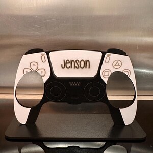 May include: A white and black wooden model of a video game controller with the name "Jenson" engraved on the top.