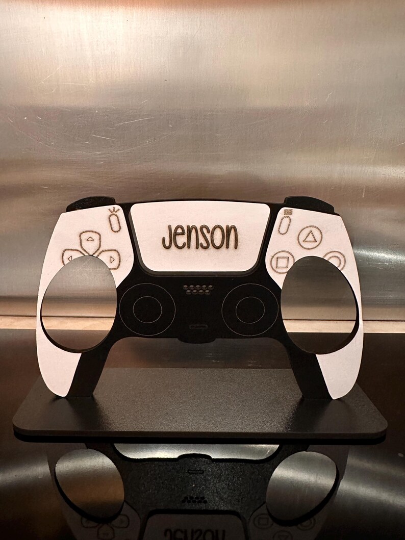 May include: A white and black wooden model of a video game controller with the name "Jenson" engraved on the top.