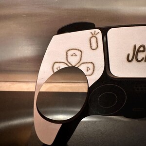 May include: A white wooden controller with black accents. The controller has a button layout similar to a Playstation 5 controller. The controller has a small, white, laser-engraved design of a flower with three petals. The controller has the name "Jen" engraved on the side.