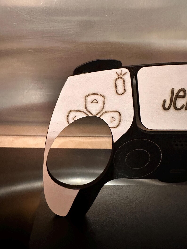 May include: A white wooden controller with black accents. The controller has a button layout similar to a Playstation 5 controller. The controller has a small, white, laser-engraved design of a flower with three petals. The controller has the name "Jen" engraved on the side.