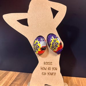 May include: A wooden Easter egg holder shaped like a person, holding two Creme Eggs. The holder has the text "Reece how do you eat yours?" on the front. The eggs are wrapped in purple and yellow foil.