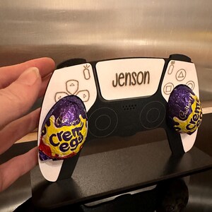 May include: A white and black PlayStation controller with the name "Jenson" engraved on the top. The controller is decorated with two purple and gold foil-wrapped chocolate eggs.