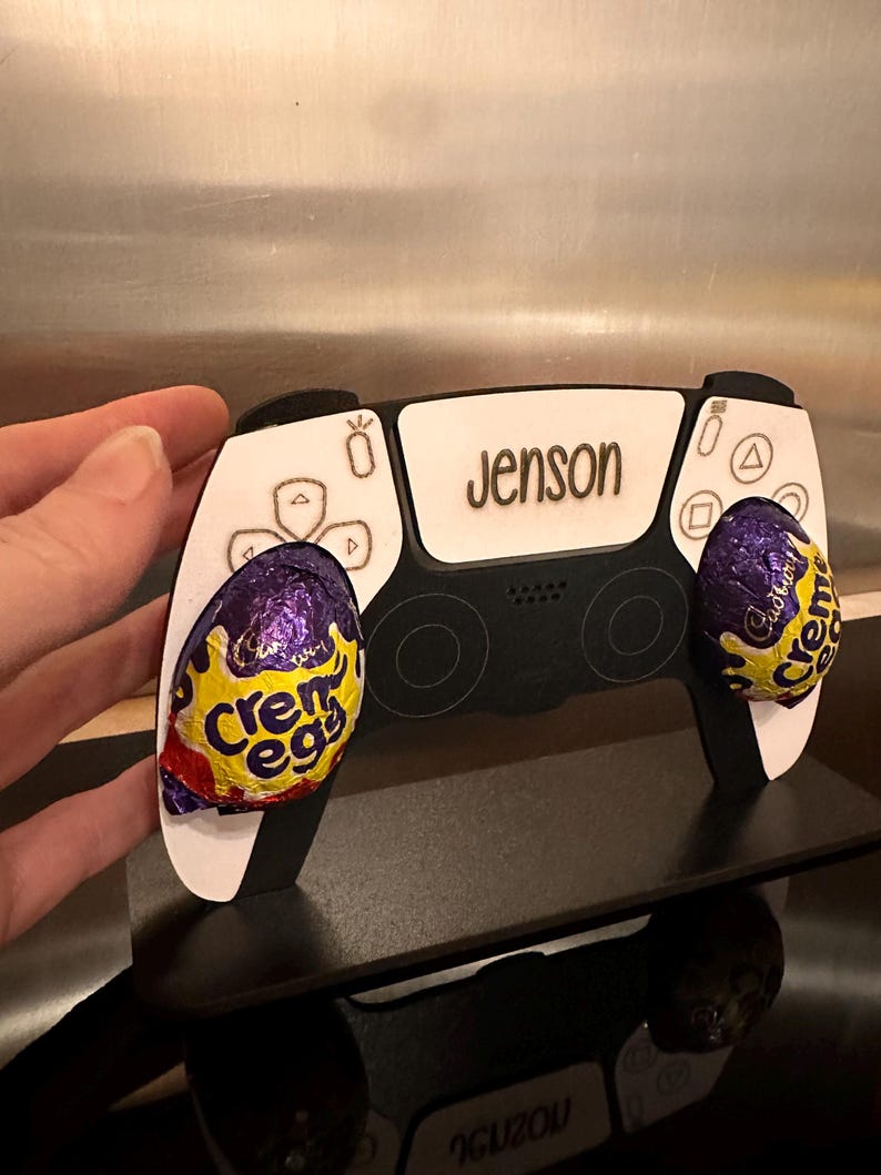May include: A white and black PlayStation controller with the name "Jenson" engraved on the top. The controller is decorated with two purple and gold foil-wrapped chocolate eggs.