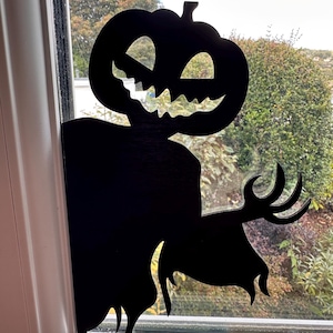 May include: Black silhouette of a Halloween pumpkin-headed scarecrow decoration. The decoration is placed in a window, with the pumpkin head and arms visible. The scarecrow has a carved, smiling face and outstretched arms.