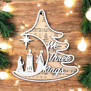 May include: A wooden Christmas tree decoration in the shape of a tree, with the words "We Three Kings" cut out. It features a star and silhouettes of the three kings. The background shows sheet music. The decoration is on a wooden surface.