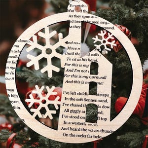 May include: Wooden Christmas decoration with a circular frame, snowflake cutouts, and a house-shaped design. The ornament has text from a poem. It is hanging on a Christmas tree.