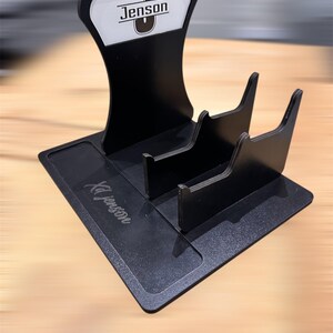 May include: Black acrylic business card holder with a stand. The stand has a white label with the name "Jenson" and a letter "J" in a circle. The base of the holder has the name "KJ Jenson" engraved in black.