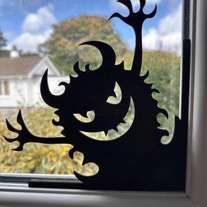 May include: A black silhouette of a cartoon monster with a toothy grin, horns, and clawed hands. The decoration is placed on a window, with a view of trees and a cloudy sky in the background. A Halloween-themed decoration.