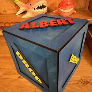 May include: A blue wooden crate with the word "DROP" printed in yellow on the side. The crate has a black trim and is decorated with the name "ALBERT" in red letters on the top. Two pink ceramic sharks are sitting on a shelf above the crate.