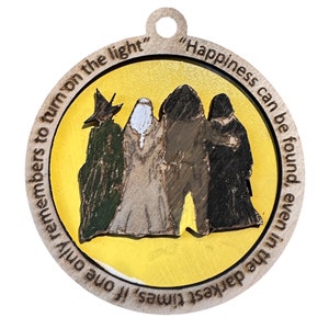 May include: A wooden ornament with a yellow background and a quote that reads "Happiness can be found, even in the darkest times if one only remembers to turn on the light". The ornament features a depiction of four figures, three of which are wearing robes and one wearing a pointed hat.