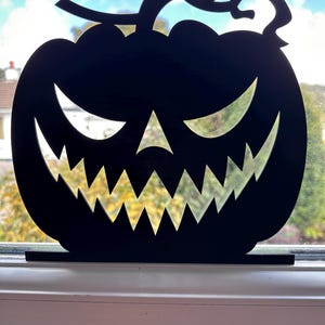 May include: A black, laser-cut Halloween pumpkin decoration with a carved face. The pumpkin has a stem and sits on a rectangular base. The pumpkin's face features triangular eyes, a triangular nose, and a jagged, toothy grin. The background shows a window with a view of trees.