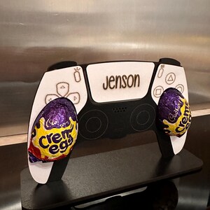 May include: A white and black PlayStation 5 controller stand with the name "Jenson" engraved on the top. Two purple and yellow Creme Eggs are placed in the controller's hand grips.