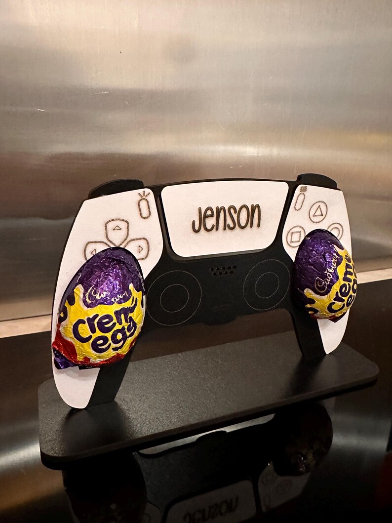 May include: A white and black PlayStation 5 controller stand with the name "Jenson" engraved on the top. Two purple and yellow Creme Eggs are placed in the controller's hand grips.