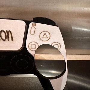 May include: A black and white wooden cutout of a PlayStation 5 controller with the word "on" written on the top left corner. The controller has a white background with black buttons and a black border.