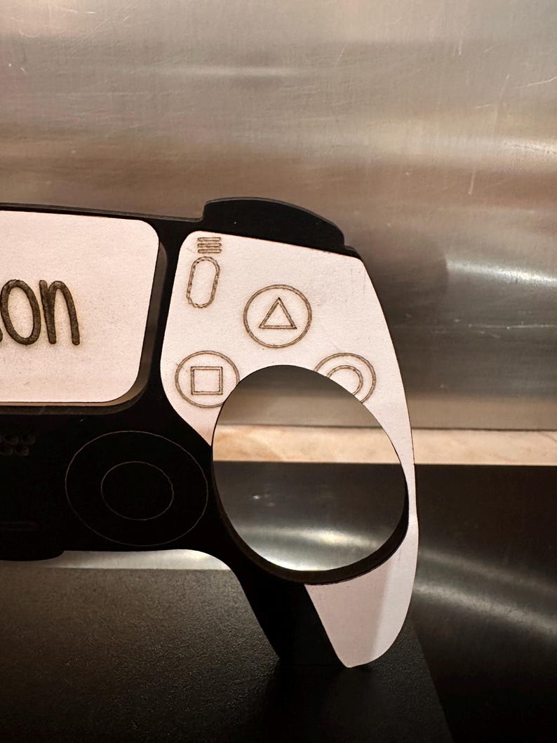 May include: A black and white wooden cutout of a PlayStation 5 controller with the word "on" written on the top left corner. The controller has a white background with black buttons and a black border.