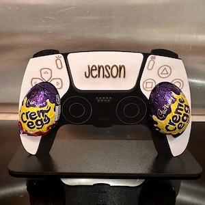 Könnte beinhalten: Ein weiß-schwarzer PlayStation 5-Controller mit dem personalisierten Namen "Jenson" auf der Vorderseite. Der Controller ist mit zwei lila und gelben Cadbury Creme Egg-Schokoladeneiern verziert.