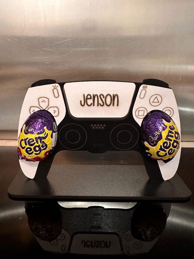 May include: A white and black PlayStation 5 controller with a personalised name "Jenson" on the front. The controller is decorated with two purple and yellow Cadbury Creme Egg chocolate eggs.