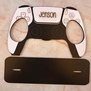 May include: A white and black PlayStation 5 controller with the name "Jenson" engraved on the top. The controller is sitting on a black stand.