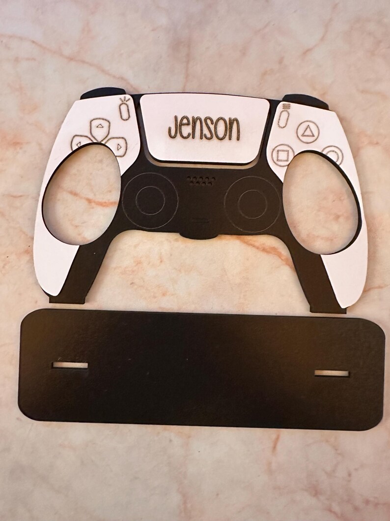 May include: A white and black PlayStation 5 controller with the name "Jenson" engraved on the top. The controller is sitting on a black stand.