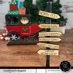 May include: A wooden signpost with six directional signs pointing to different areas of the North Pole. The signs read: North Pole, Toy Factory, Sleigh Parking, Reindeer Games, Candy Cane Lane, and Santa's Workshop.