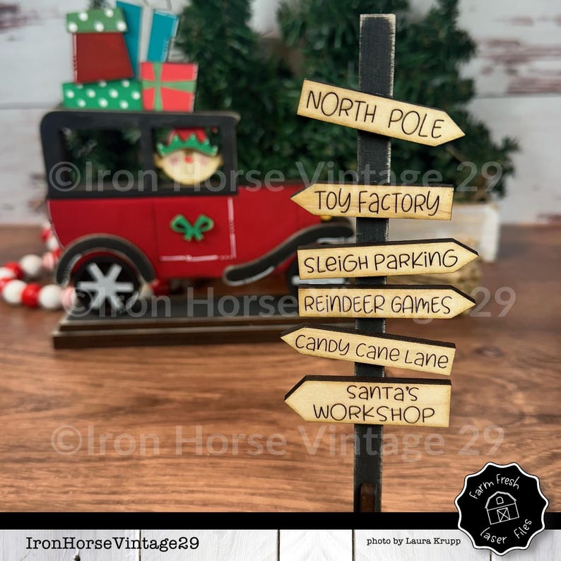 May include: A wooden signpost with six directional signs pointing to different areas of the North Pole. The signs read: North Pole, Toy Factory, Sleigh Parking, Reindeer Games, Candy Cane Lane, and Santa's Workshop.