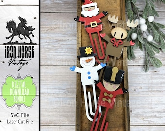 Op de afbeelding: Houten kerstthema paperclips met de kerstman, een sneeuwpop, een rendier en een soldaat. De clips zijn rood, wit, bruin en zwart, met gele en blauwe accenten. De afbeelding bevat ook de tekst "DIGITAL DOWNLOAD BUNDLE" en "SVG File Laser Cut File."