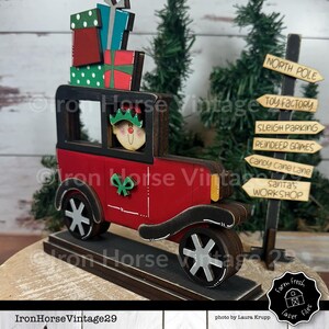 May include: A wooden Christmas decoration of a red vintage car carrying a stack of wrapped gifts and a smiling elf. The car is on a wooden base and there is a wooden signpost with the words "North Pole", "Toy Factory", "Sleigh Parking", "Reindeer Games", "Candy Cane Lane", and "Santa's Workshop" on it.