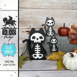 May include: Three black wooden cat skeleton cutouts with white painted details. The largest cutout is in the center, the smallest is on the right, and the middle-sized cutout is on the left. The cutouts are on a wooden surface with fall leaves and pumpkins.